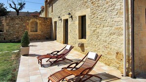A stone building with a tiled patio, two wooden lounge chairs, and a potted plant. - La Grange Des Endrevies (Sarlat La Caneda)