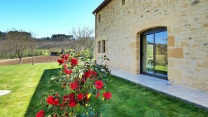 A stone building with a glass door and windows, surrounded by a well-maintained lawn and a bush of red flowers. - La Grange Des Endrevies (Sarlat La Caneda)