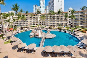 A resort pool with lounge chairs and umbrellas, surrounded by palm trees and hotel buildings. - Villa del Palmar PV by Villa TS Rentals (Puerto Vallarta)