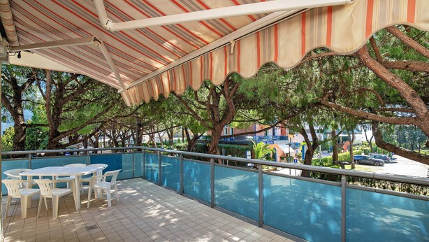 A covered outdoor seating area with tables and chairs, a railing, and trees in the background. - Apartment 'Condominio Lara A16' with Private Terrace and Air Conditioning (Jesolo)