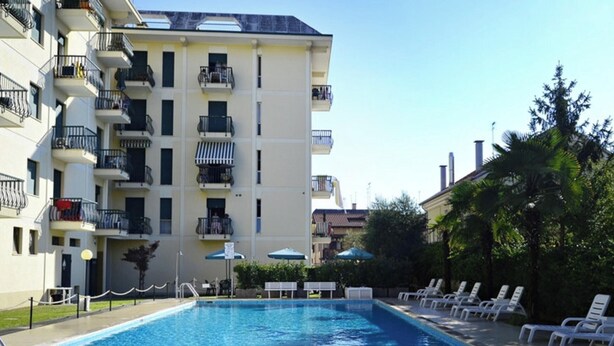 A swimming pool with lounge chairs, a building with balconies, and a clear sky. - Apartment 'Residence Rubino Benelux 01' with Shared Pool, Terrace, and Air Conditioning (Jesolo)