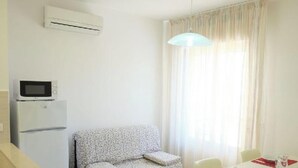 A room with a white air conditioner, a flat-screen TV on a white refrigerator, a floral sofa, a dining table with chairs, and a large window with sheer curtains. - Apartment 'Residence Rubino Benelux 01' with Shared Pool, Terrace, and Air Conditioning (Jesolo)