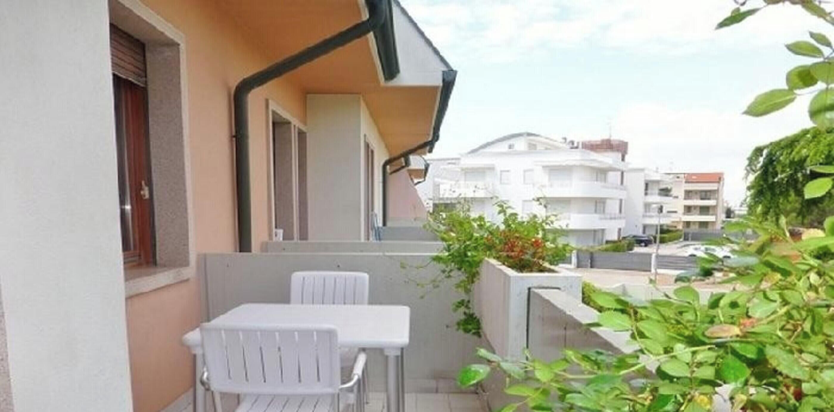 Un balcón con una mesa y sillas, una jardinera de concreto con plantas y vista a edificios cercanos.