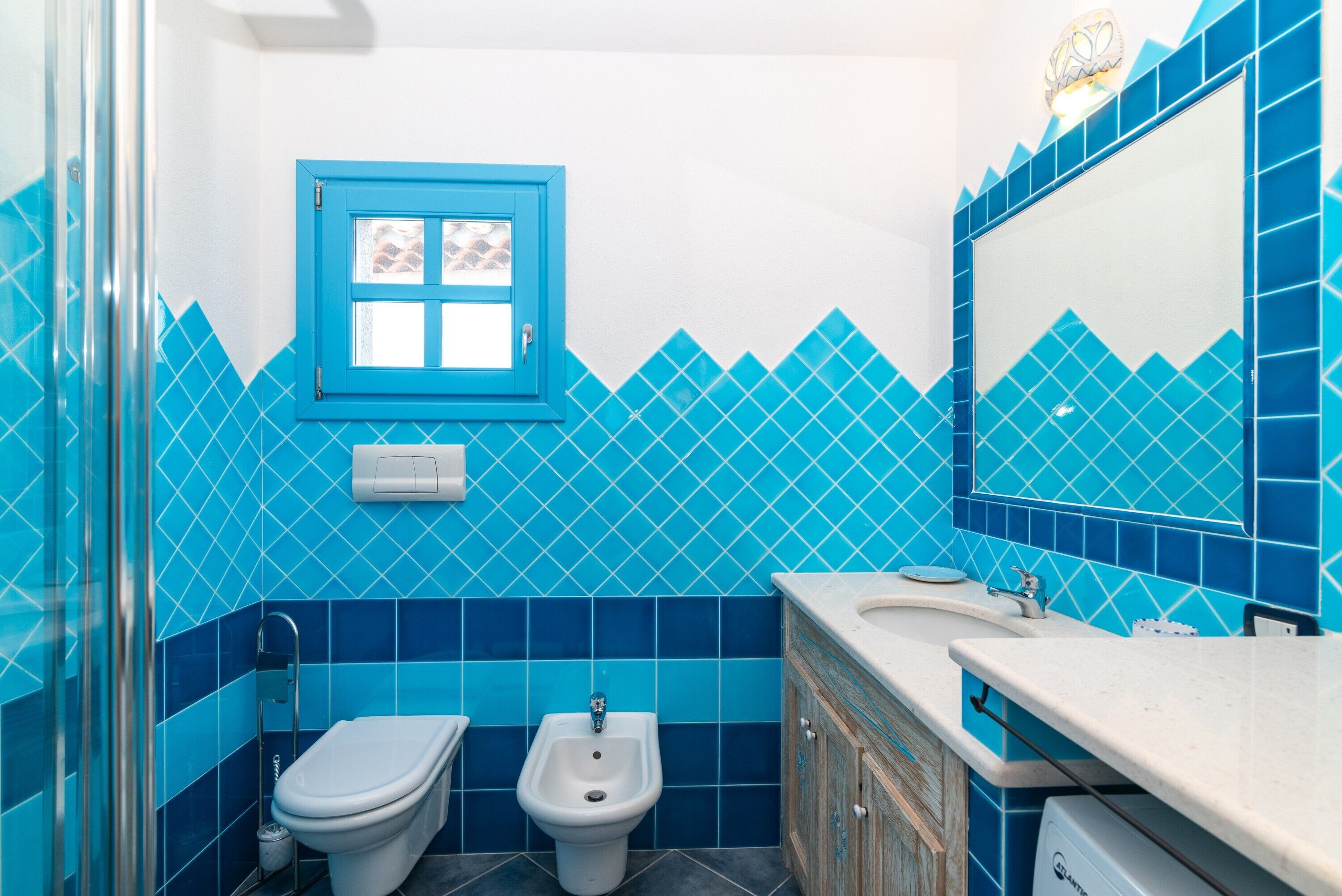 A bathroom with a blue-tiled wall, a white toilet, a bidet, a sink with a mirror, and a cabinet.