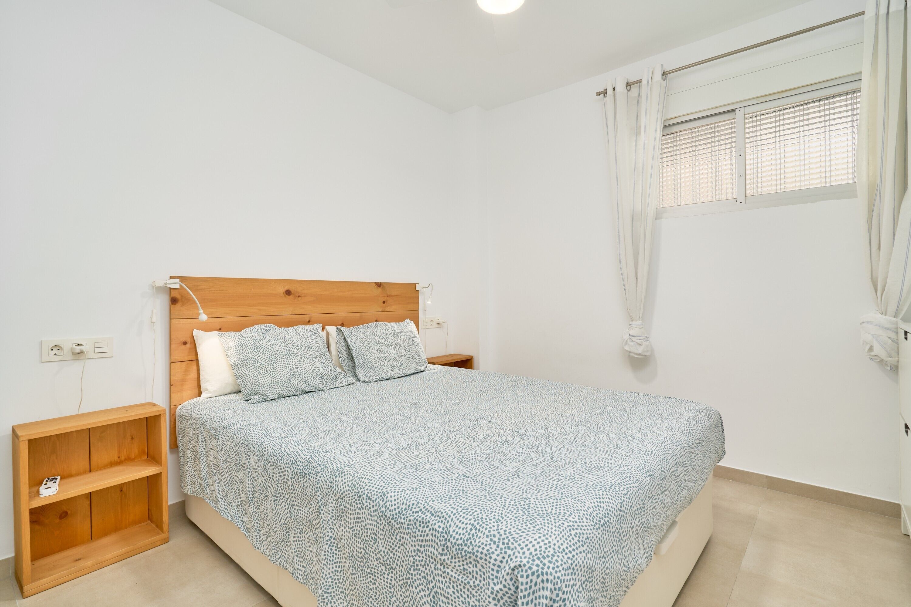 Un dormitorio con cabecera de madera, cama con ropa de cama estampada, mesita de noche, estante de madera y ventana con cortinas.