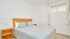 A bedroom with a wooden headboard, a bed with patterned bedding, a nightstand, a wooden shelf, and a window with curtains. - Apartment 'Apartamento Moderno' with Sea View, Wi-Fi and Air Conditioning (Almuñecar)