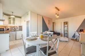 Eine moderne Küche mit weißen Schränken, einem Esstisch mit Platzdeckchen und einem Wohnbereich mit Sofa und Bücherregal.