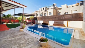 A swimming pool with a wooden deck, a covered seating area, and a building with a flat roof in the background. - Holiday Home 'Goleta' with Private Pool, Wi-Fi and Air Conditioning (El Puerto de Santa María)