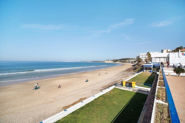 A beach with a clear sky, a sandy shore, and a few people. There are buildings in the background.