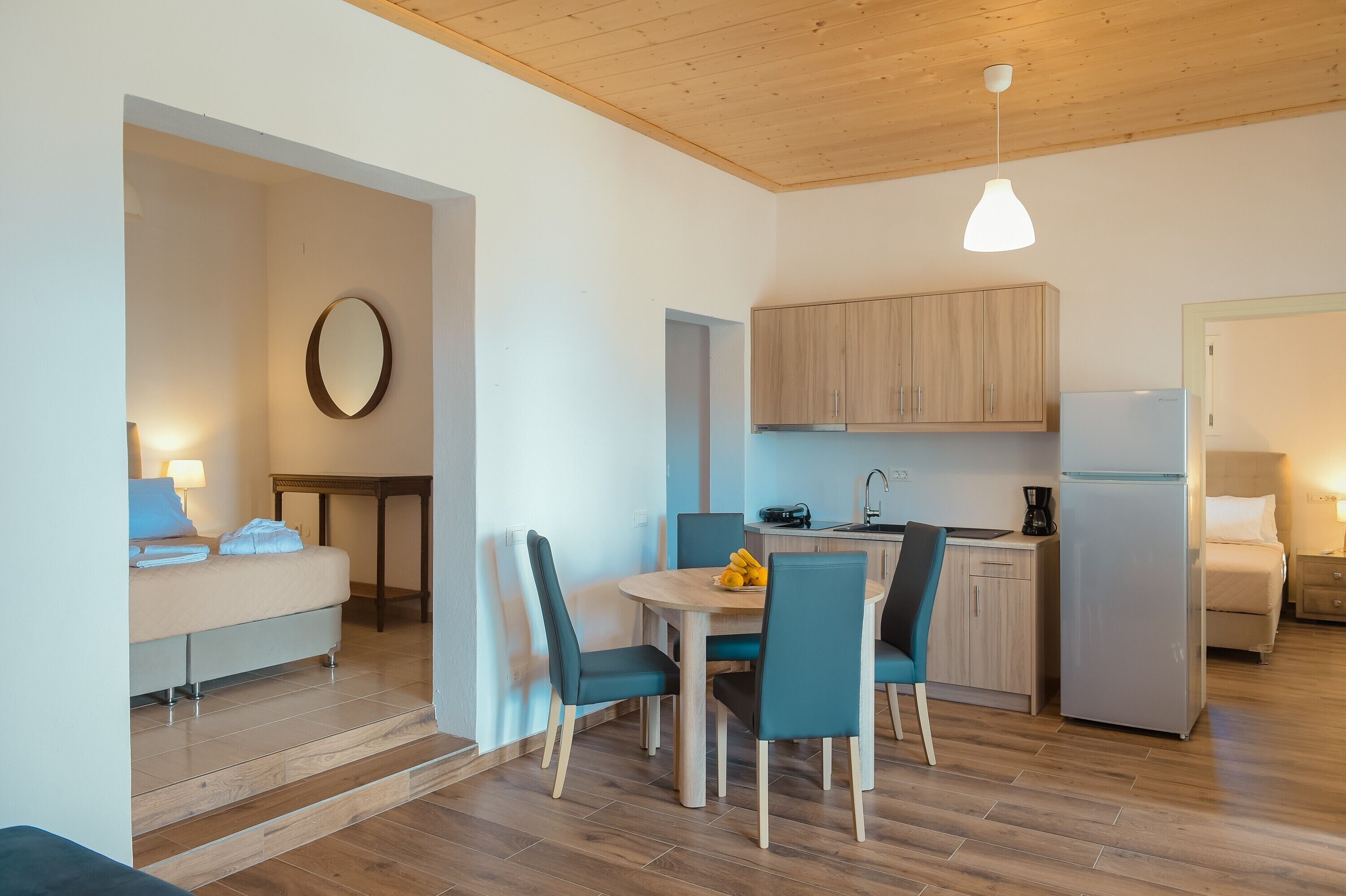 Une chambre d’hôtel compacte comprenant un lit, un petit coin repas avec une table et des chaises, une cuisine équipée d’armoires et d’un réfrigérateur, et un plafond en bois.