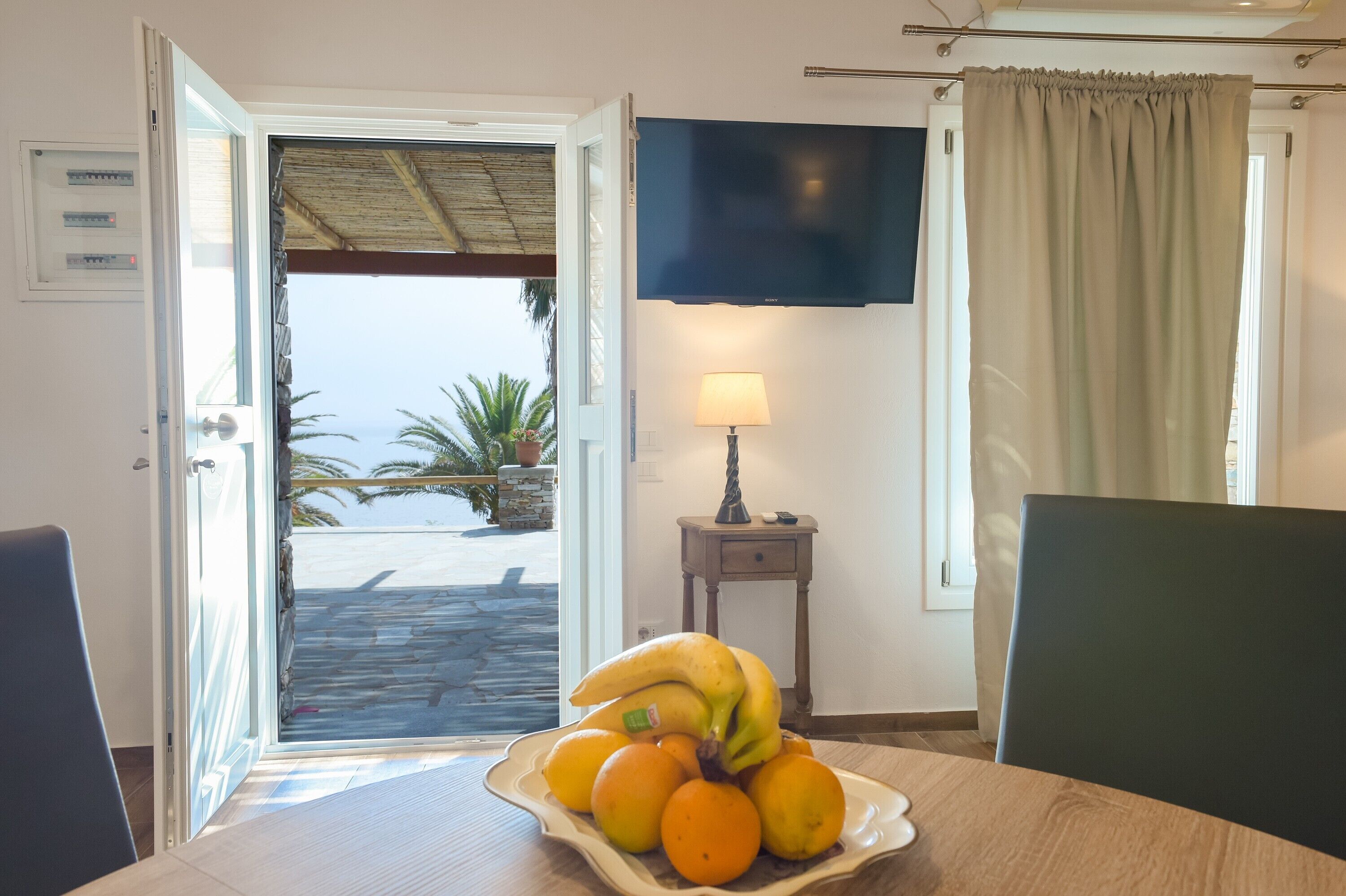 Une pièce comprenant une table à manger, un panier de fruits contenant des bananes et des oranges, un téléviseur fixé au mur, une lampe et une vue sur l’extérieur.
