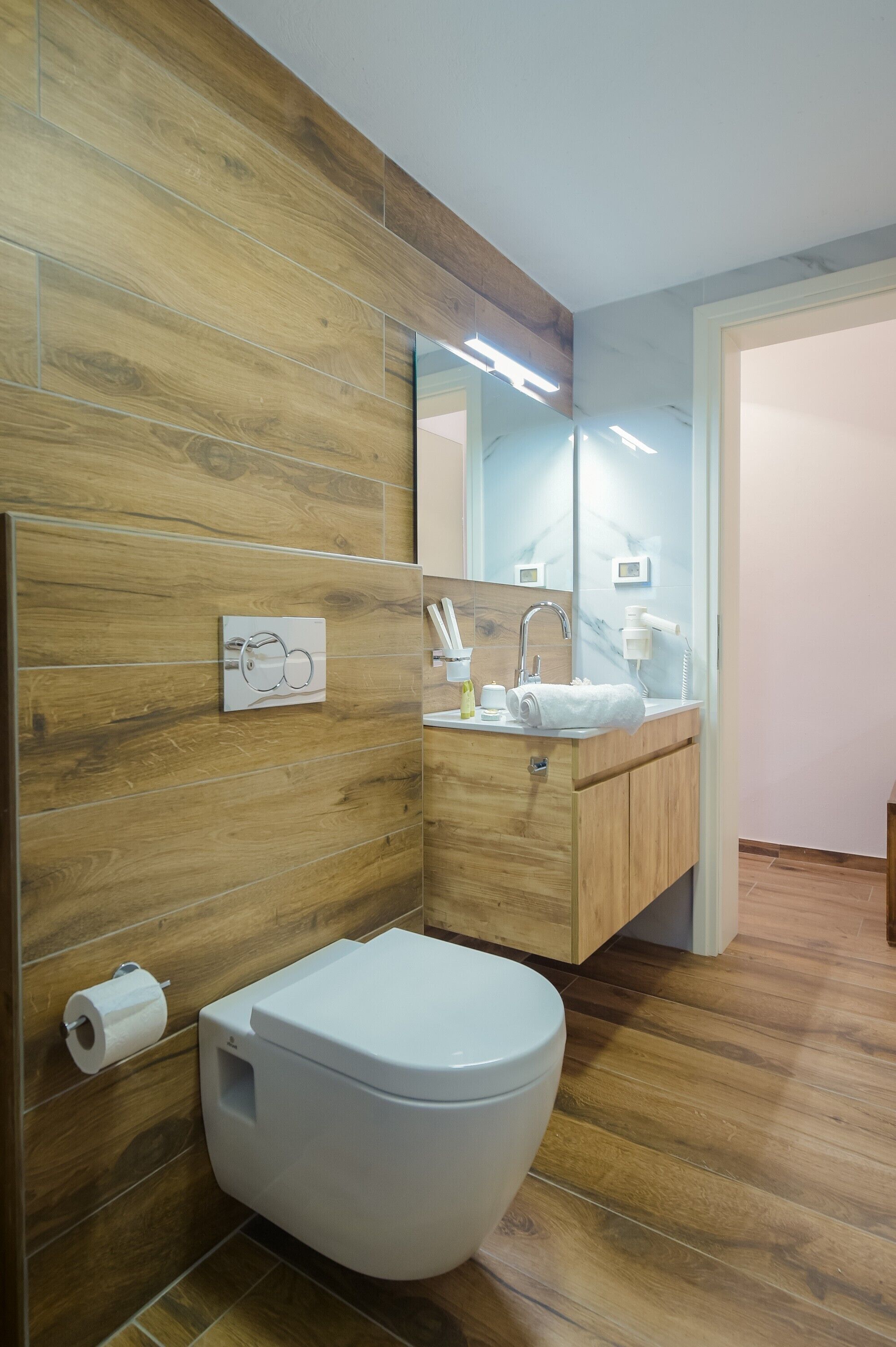 Un baño moderno con pared de madera, inodoro blanco, lavamanos con espejo y un perchero para toallas.