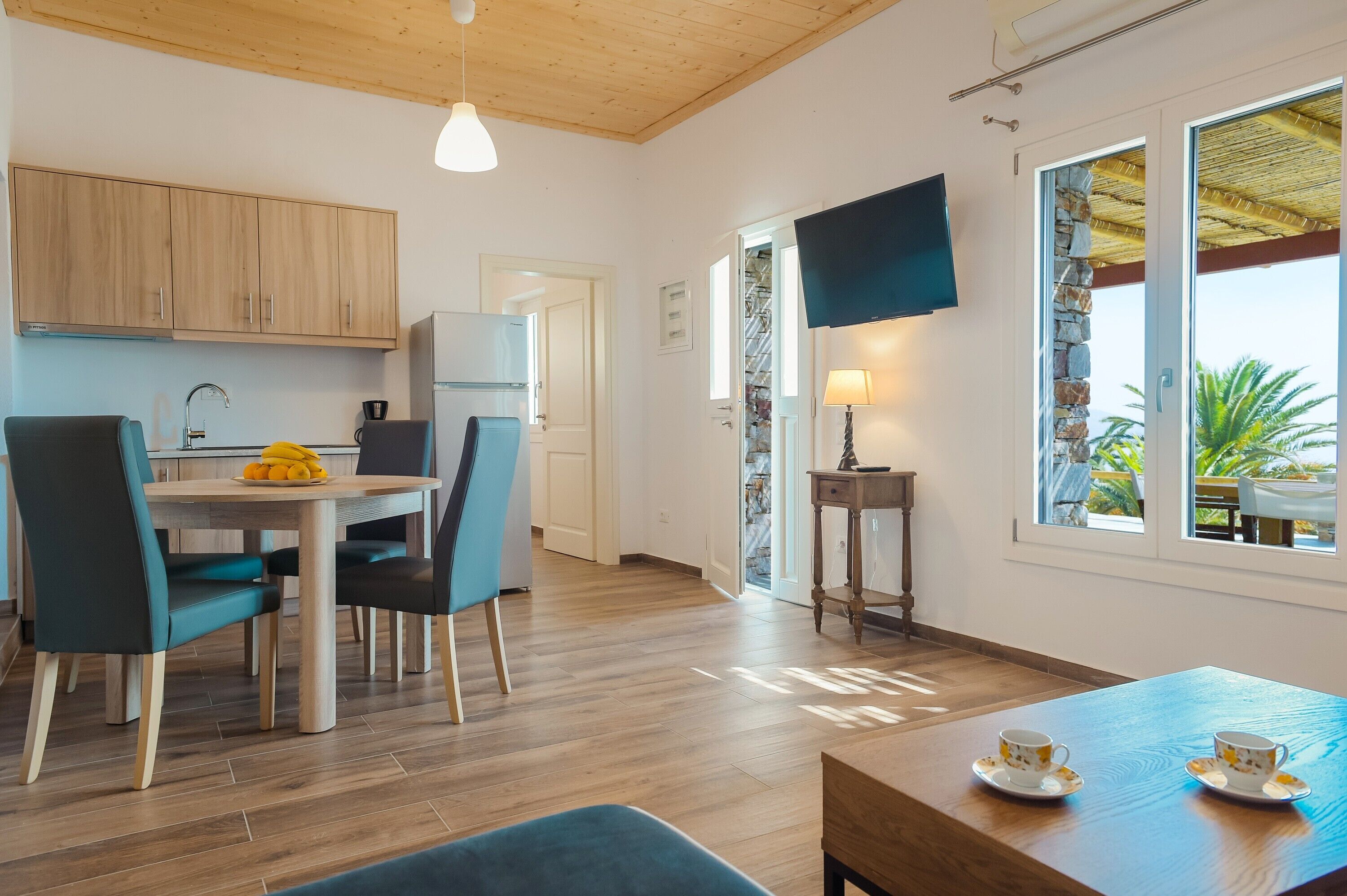 Une cuisine moderne avec des meubles en bois, une table à manger avec des chaises, un téléviseur fixé au mur et une fenêtre donnant sur un paysage verdoyant.