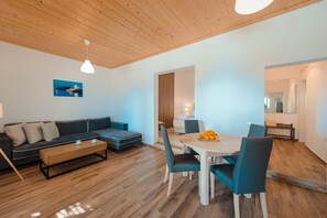A modern living room with a sofa, coffee table, dining table with chairs, wooden flooring, and a ceiling with wooden beams.