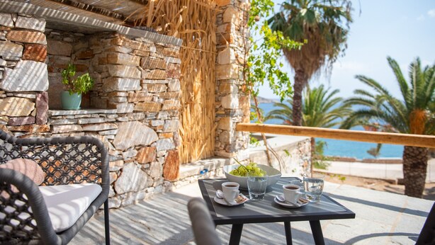 Un mur de pierre surmonté d’un toit en osier tressé, une chaise en rotin, une table avec un panier de fruits et une vue sur la mer.