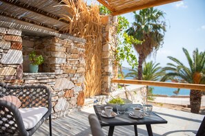 Un mur de pierre surmonté d’un toit en osier tressé, une chaise en rotin, une table avec un panier de fruits et une vue sur la mer.