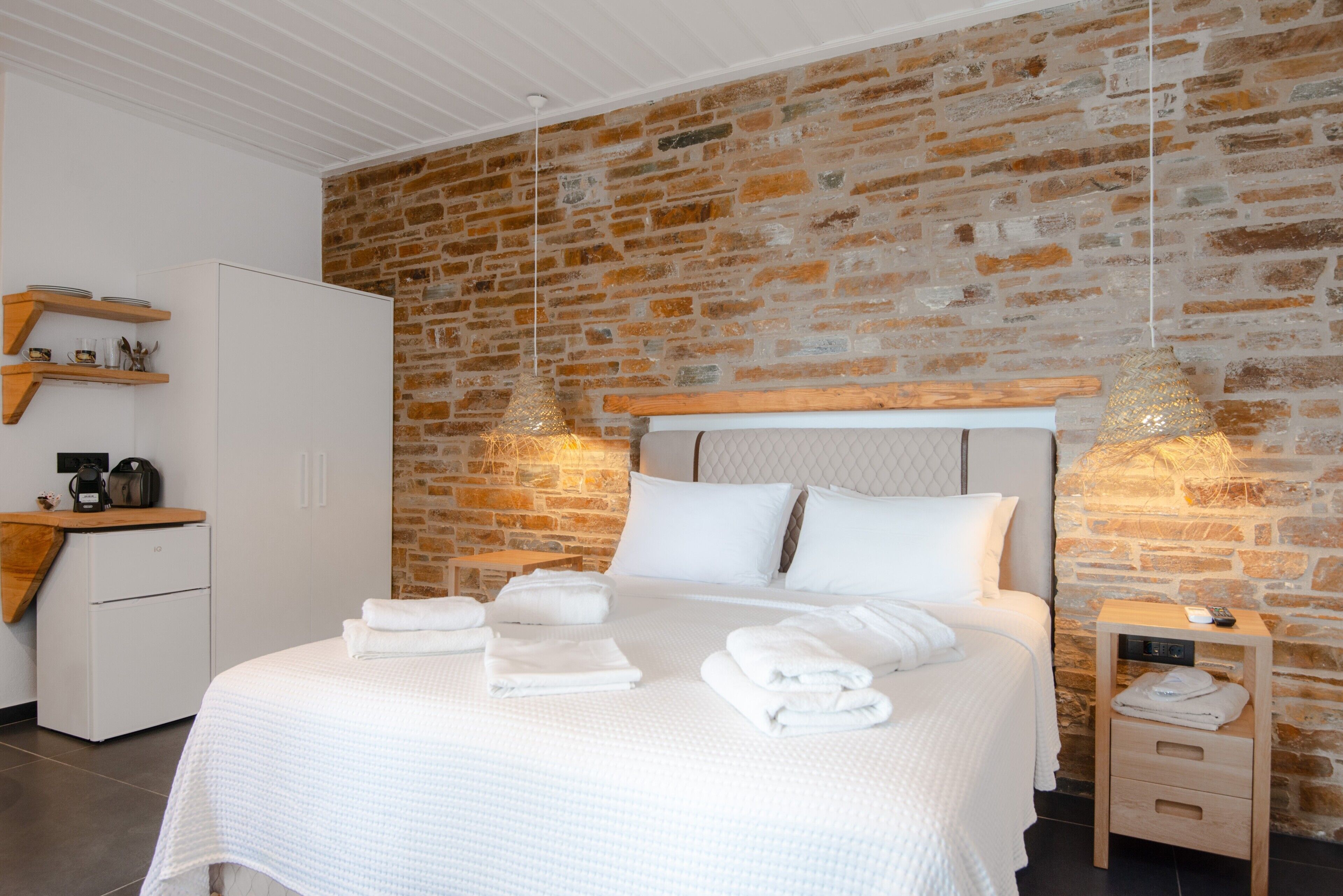 Moderni hotellihuone, jossa on tiiliseinä, siististi pedattu sänky, pieni työpöytä ja yöpöytä, jossa on lamppu.