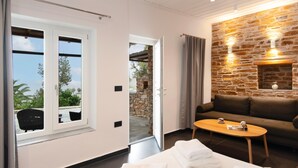 A room with a bed, a wooden table, a brown sofa, a stone wall, and a view through a window. - White & Black Suites – Suite Three (Platys Gialos)