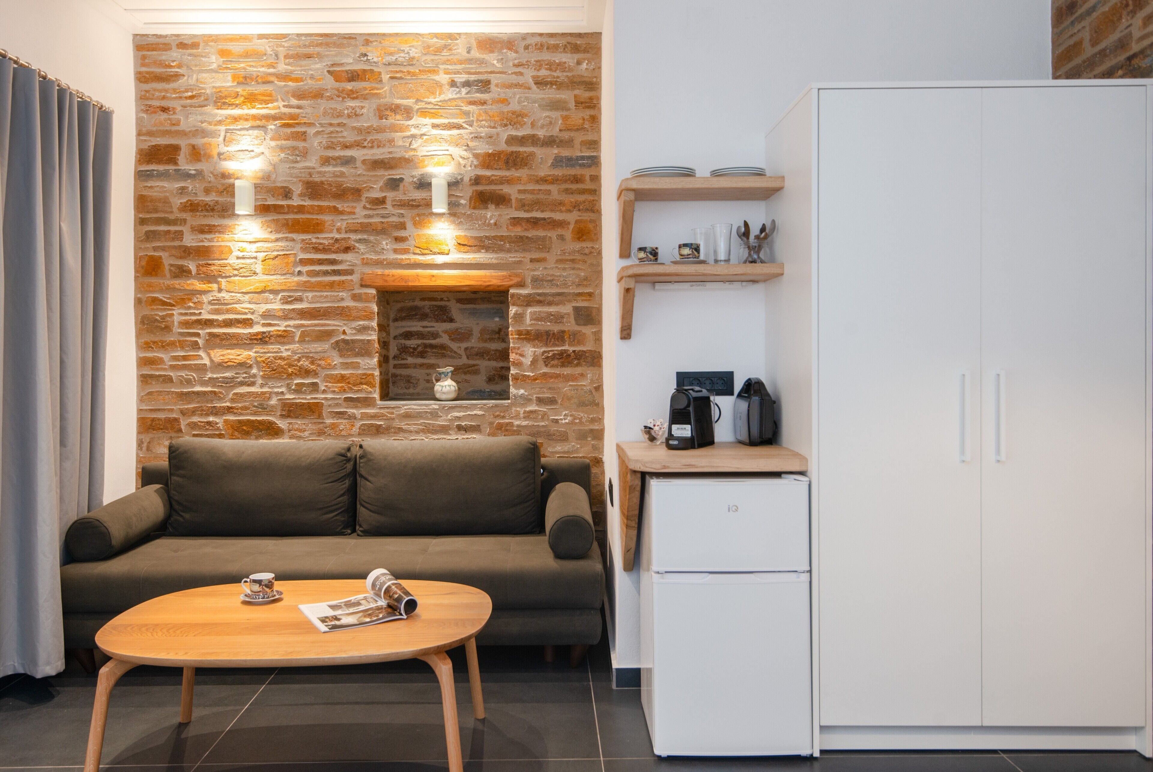 Un espace de vie moderne avec un mur en pierre, un canapé, une table basse, un réfrigérateur blanc et une étagère en bois où sont rangés des ustensiles de cuisine.