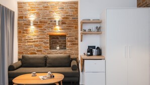 A modern living area with a stone wall, a sofa, a coffee table, a white refrigerator, and a wooden shelf with kitchenware. - White & Black Suites – Suite Three (Platys Gialos)