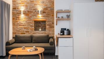 Un espace de vie moderne avec un mur en pierre, un canapé, une table basse, un réfrigérateur blanc et une étagère en bois où sont rangés des ustensiles de cuisine.