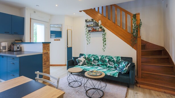 A modern kitchen with blue cabinets, a wooden staircase, and a green sofa with a tropical print. - Villa 'Oasis Tropicale A Cleguerec' with Private Terrace, Private Garden and Wi-Fi (CLEGUEREC)