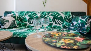 A room with a green sofa, two clear glasses on a wooden table, and a decorative plate with tropical flowers. - Villa 'Oasis Tropicale A Cleguerec' with Private Terrace, Private Garden and Wi-Fi (CLEGUEREC)