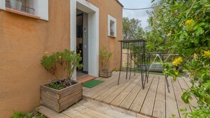 Una terrassa de fusta amb plantes en testos, una pèrgola metàl·lica i l'entrada d'un edifici.