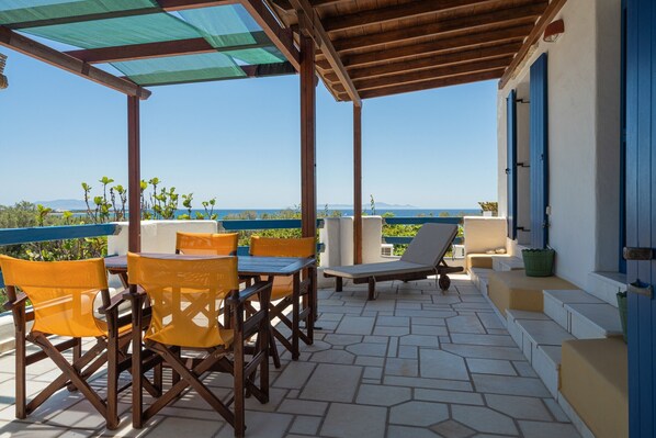 A covered outdoor seating area with a view of the sea, wooden furniture, and a stone floor.