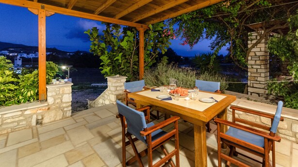 An outdoor dining area with a wooden table and chairs, stone walls, and a view of a distant town at dusk. - Vacation Home "Maria's Village Two Bedroom House 5" with Sea View, Terrace & Wi-Fi (Paros)