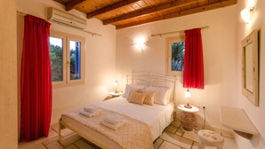 A bedroom with a wooden ceiling, a bed with white linens, two bedside tables with lamps, and a window with red curtains. - Vacation Home "Maria's Village Two Bedroom House 5" with Sea View, Terrace & Wi-Fi (Paros)