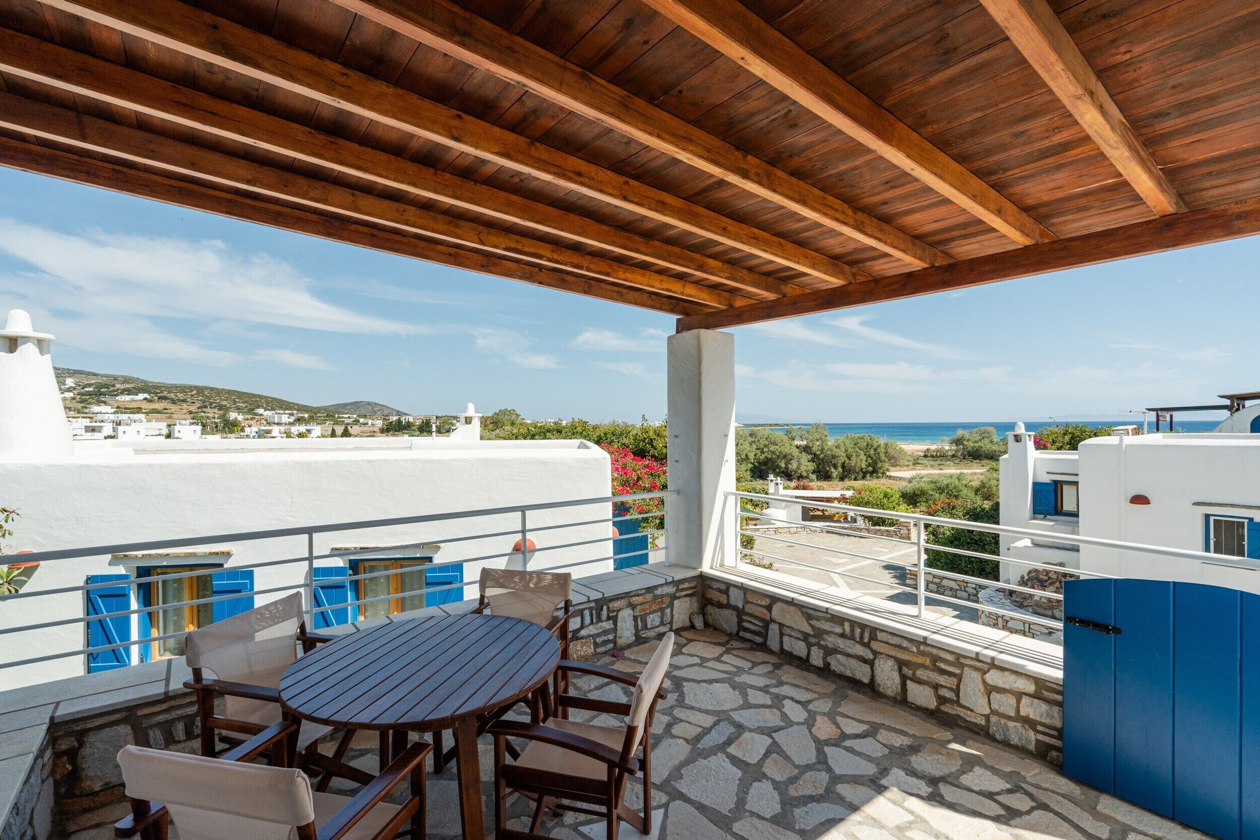 Eine überdachte Terrasse im Freien mit Tisch und Stühlen, einem Steinboden und Blick auf das Meer und Gebäude in der Ferne.