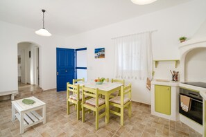 Un comedor con una mesa y sillas, una mesita auxiliar, una puerta azul, una cocina con estufa y una ventana con cortinas.