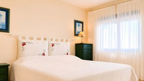 Una cama bien hecha con cubrecama blanco y almohadas florales, una mesita de noche verde, una pequeña mesa con lámpara y una ventana con cortinas traslúcidas.