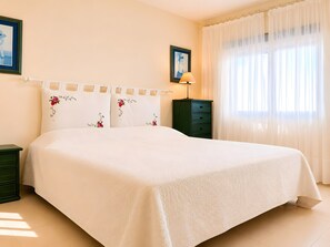 Ein ordentlich bezogenes Bett mit weißer Bettdecke und blumigen Kissen, ein grüner Nachttisch, ein kleiner Tisch mit Lampe und ein Fenster mit durchscheinenden Vorhängen.