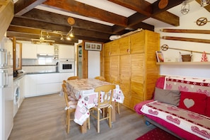 Ein gemütliches Zimmer mit Holzbalkendecke, einem Esstisch mit Stühlen, einer Couch mit herzenmusternder Bettwäsche und einer Küche im Hintergrund.