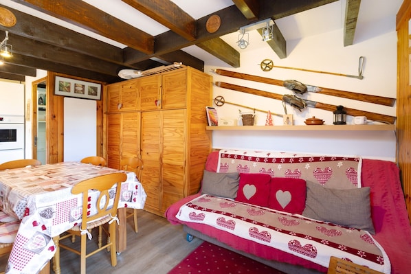 Ein gemütliches Zimmer mit einem Holzschrank, einer Couch mit Kissen in Herzmuster, einem Esstisch mit kariertem Tischläufer und Ski sowie Skistöcken an der Wand.