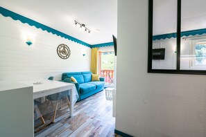 A room with a blue sofa, a white table, a clock on the wall, and a window with a view of the outside. - Apartment 'Appartement 4-6 Personnes' with Mountain View, Balcony and Wi-Fi (Saint-François-Longchamp)