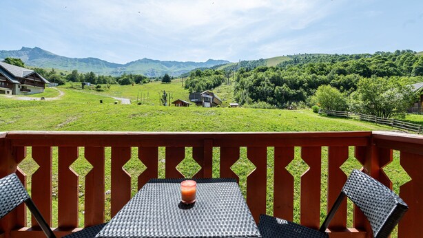 Un balcon avec une table et des chaises, un verre à la main, et une vue imprenable sur des collines verdoyantes et des montagnes.