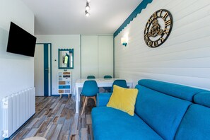 A room with a blue sofa, a dining table with chairs, a wall-mounted clock, and a television on the wall. - Apartment 'Appartement 4-6 Personnes' with Mountain View, Balcony and Wi-Fi (Saint-François-Longchamp)