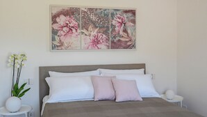 A bedroom with a bed, white and pink pillows, a bedside table, a nightstand, and a wall-mounted floral artwork. - Appartamento Alba - Le 3 Rose Apartments (Sicilia)