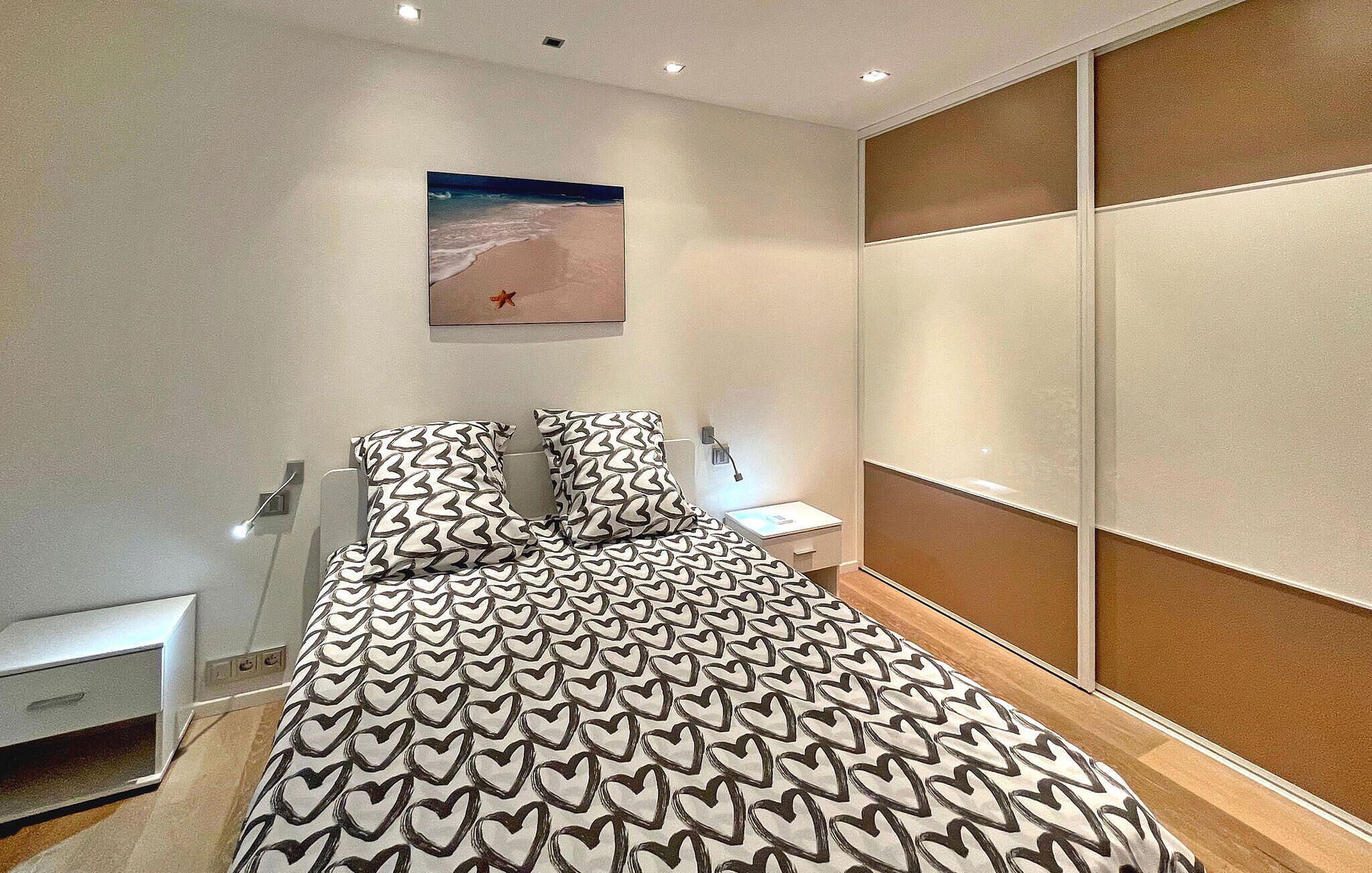 Un dormitorio moderno con una colcha estampada, una mesita de noche, una lámpara de noche y una obra de arte colgada en la pared que representa una escena de playa.
