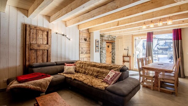A cozy living room with a large sectional sofa, wooden beams on the ceiling, a stone wall, and a view of the outside. - LA Maison Bleue - Bessans (BESSANS)