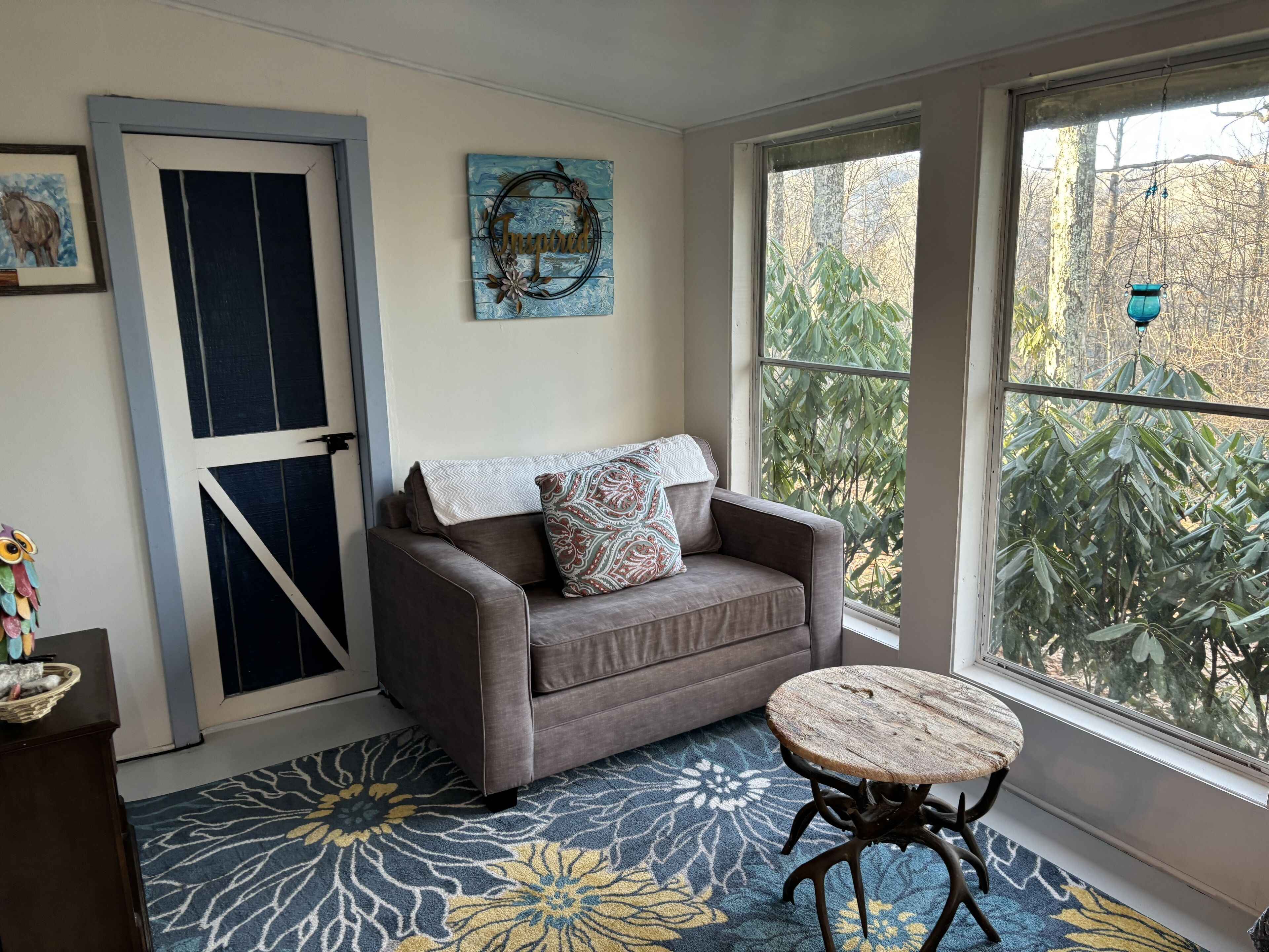 A cozy room with a gray sofa, a small round table, a blue patterned rug, a decorative wall piece, and a view of trees through a window.