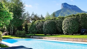 A swimming pool with lounge chairs, surrounded by well-maintained hedges and trees, with a mountain in the background.