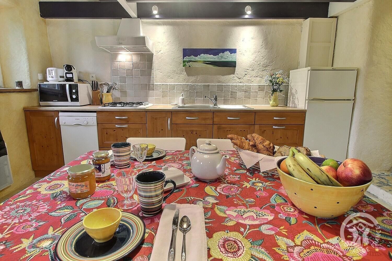 Una cocina con armarios de madera, microondas, lavamanos y una mesa de comedor con mantel floral, platos y un cuenco de fruta.