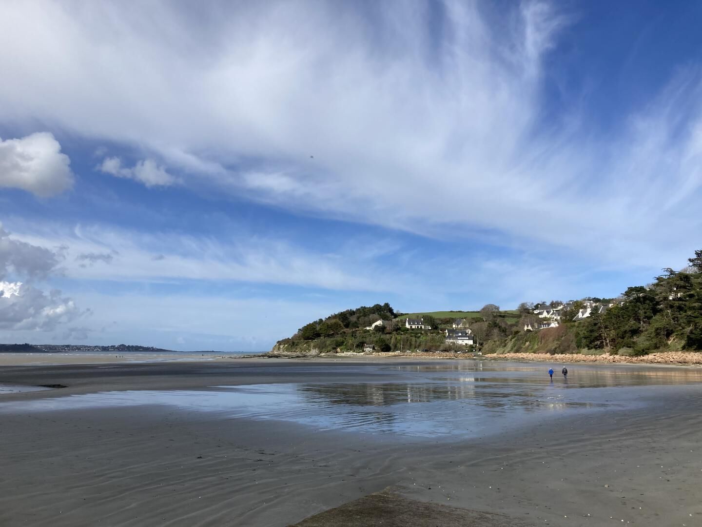 Ein Strand mit ruhiger Wasserfläche, blauem Himmel und Häusern, die an einem Hügel im Hintergrund liegen.