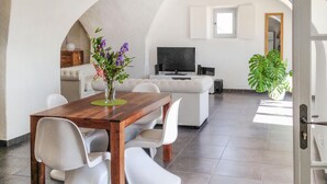 A modern living room with a wooden dining table, white chairs, a sofa, a TV, and a potted plant.