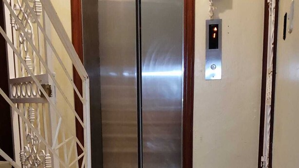 En trappe fører op til en elevator med dobbelt døre, med gelænder på venstre side og en vægmonteret lyskontakt på højre side.