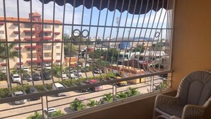 En balkon med udsigt til en parkeringsplads og bygninger. To fletstole og et lille rundt bord med en potteplante.